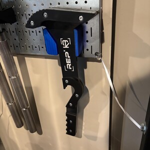 May include: Black and blue weightlifting tool with the text "REP x K2" attached to a metal pegboard. The tool has a hook and a serrated edge. Other weightlifting equipment is visible in the background.