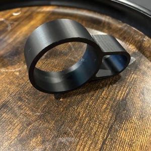 May include: A black metal ring with a smaller, attached loop. The ring and loop are made of a solid, matte black material. The object is set against a wooden surface.