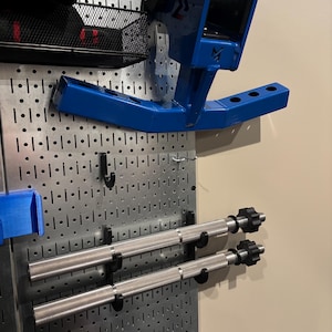 May include: A blue metal weightlifting rack attachment with a black handle and a logo. Two silver barbell handles are mounted below. The items are attached to a metal pegboard.