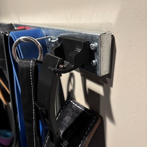 May include: A metal rack mounted on a beige wall, holding various belts and straps. The rack is made of silver metal and has a black hook. Several belts of different colors and materials hang from the rack, including blue, black, and white.