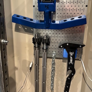 May include: A collection of weightlifting equipment, including a blue T-bar row attachment, two silver barbell handles, a chain, and a black REP Fitness tool. The items are mounted on a metal pegboard.