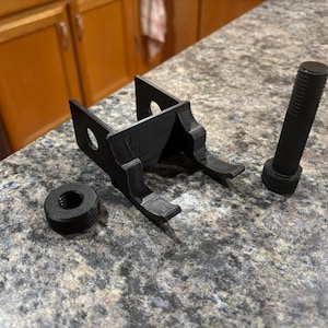 May include: Black 3D-printed hardware components, including a bracket with two holes, a bolt, and a nut. The parts are arranged on a speckled countertop, showcasing a functional design.