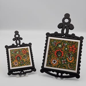 May include: Two black cast iron trivets with floral tile inserts. The tiles feature a repeating pattern of orange, white, and green flowers on a gold background. The trivets have decorative scrollwork and a loop for hanging.