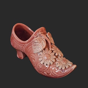 May include: A decorative, ceramic shoe in a dusty rose color. The shoe features embossed floral and leaf details, including three prominent daisy-like flowers on the toe. The heel is small and curved, and the shoe has a pointed toe.