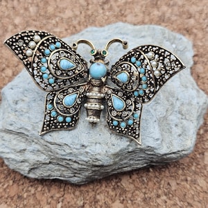 May include: A detailed butterfly brooch with a vintage aesthetic. The wings are adorned with turquoise-colored stones, faux pearls, and clear crystals, set in a bronze-toned metal filigree. The butterfly's body features a large turquoise stone and small faux pearls.