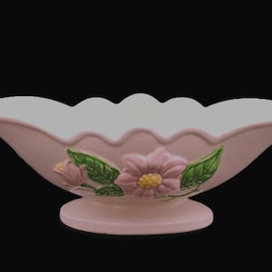 May include: A light pink ceramic flower vase with a scalloped rim and a pedestal base. The vase is decorated with pink flowers and green leaves. The vase is a decorative item.