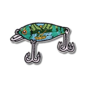 Puede incluir: Parche bordado de señuelo de pesca con una escena de paisaje detallada en el interior. El señuelo es verde azulado y blanco, con un lago, árboles y montañas. Dos anzuelos plateados cuelgan debajo. Un parche único para los entusiastas de la pesca.