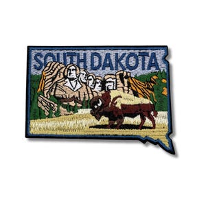 May include: Embroidered patch in the shape of South Dakota, featuring Mount Rushmore and a bison. The text "SOUTH DAKOTA" is embroidered across the top in blue. The patch has a blue border and is on a white background.