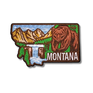 Puede incluir: Parche bordado con la forma de Montana, con un oso pardo, montañas y una cascada. La palabra "MONTANA" se muestra en letras blancas. El parche tiene un borde marrón y un fondo blanco.