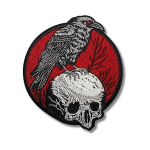 Può includere: Toppa ricamata raffigurante un corvo nero con occhi rossi appollaiato su un teschio bianco su sfondo circolare rosso. Il corvo ha piume strutturate e dettagliate. La toppa ha un bordo nero.