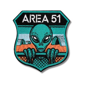 Puede incluir: Parche bordado con un alienígena del Área 51. El parche tiene forma de escudo con las palabras "AREA 51" en la parte superior. El alienígena es de color verde azulado con grandes ojos negros y está sobre un fondo verde azulado, naranja y azul.