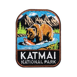 May include: Embroidered patch featuring a brown bear catching a salmon in a river, with mountains and trees in the background. The patch has the text "KATMAI NATIONAL PARK" in white letters on a black background, with a brown border.