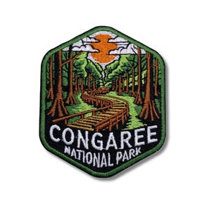 May include: Embroidered patch featuring a scenic view of Congaree National Park. The design includes a wooden walkway winding through a forest, with a setting sun and the park's name in white text.