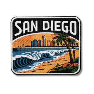 Puede incluir: Parche bordado con una escena costera y el texto "SAN DIEGO". El diseño incluye una playa, olas del océano, palmeras y un paisaje urbano contra un cielo naranja y azul.