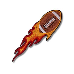 Puede incluir: Parche bordado que representa un balón de fútbol americano marrón con rayas blancas y cordones, rodeado de llamas estilizadas en tonos rojos, naranjas y amarillos. El diseño da la impresión de que el balón está en llamas.