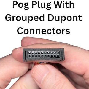 May include: A black Pog Plug with grouped Dupont connectors. The image shows a close-up of the plug, held in a person's hands. The text "Pog Plug With Grouped Dupont Connectors" is displayed at the top of the image.
