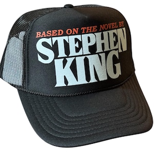 May include: Black trucker hat with the text "BASED ON THE NOVEL BY STEPHEN KING" in white and red lettering. The hat has a mesh back and a curved brim.