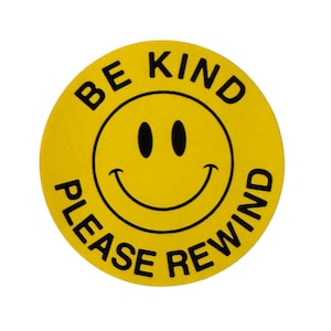 May include: A round, yellow sticker with a smiling face and the words "BE KIND PLEASE REWIND" in black, curved around the smiley face. The sticker has a simple, bold design.