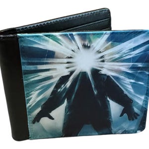 May include: Black bi-fold wallet featuring a graphic from a horror film. The wallet's exterior is black, while the interior displays a blue and white image of a figure with a bright light emanating from its head. A stylish accessory.
