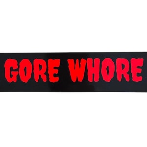 May include: A rectangular black sticker with the words "GORE WHORE" in bright red, dripping-style lettering. The sticker has a glossy finish and is set against a plain white background.