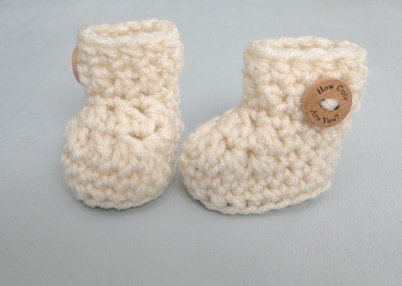 K&ouml;nnte beinhalten: Ein Paar geh&auml;kelte Babyschuhe in Cremefarbe mit einem Holzknopf, auf dem "How Cute Are You?" steht.