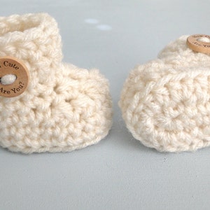 K&ouml;nnte beinhalten: Zwei wei&szlig;e H&auml;kel-Babyschuhe mit Holz-Kn&ouml;pfen. Ein Knopf sagt "How Cute Are You?"