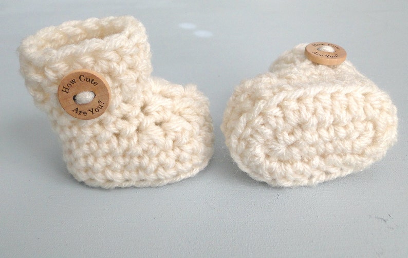 K&ouml;nnte beinhalten: Zwei wei&szlig;e H&auml;kel-Babyschuhe mit Holz-Kn&ouml;pfen. Ein Knopf sagt "How Cute Are You?"