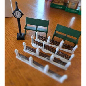 May include: Miniature park scene with a black clock tower, two green benches, and white and black decorative fences. The scene is set on a wooden surface, suggesting a model or diorama.