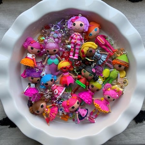 May include: A collection of colorful doll charms with various hairstyles and outfits, including dresses and skirts. The dolls are in a white, ruffled-edge dish, with some charms featuring pink, orange, and green accents.