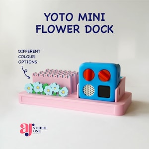 Op de afbeelding: Een roze Yoto Mini Flower Dock met een blauwe kubusvormige component met rode cirkels en een luidspreker. De dock bevat een roze bloembed met blauwe en witte bloemaccenten. Tekst op de afbeelding luidt "YOTO MINI FLOWER DOCK" en "DIFFERENT COLOUR OPTIONS".