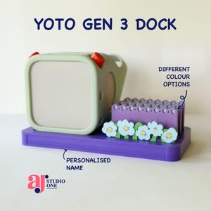 Op de afbeelding: Een Yoto Gen 3 Dock. De basis is paars, met een mintgroene luidspreker en een paarse houder versierd met lichtblauwe en witte bloemen. De tekst "YOTO GEN 3 DOCK" staat bovenaan. De tekst "DIFFERENT COLOUR OPTIONS" staat rechts. De tekst "PERSONALISED NAME" staat onderaan.