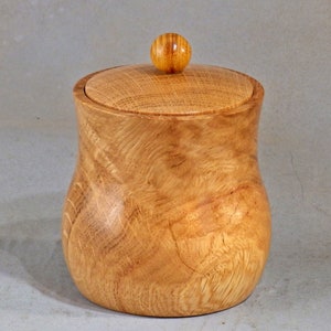 Puede incluir: Un tarro de madera con tapa y un pomo redondo en la parte superior. El tarro está hecho de madera marrón claro con un patrón de veta natural.