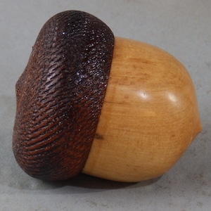 May include: A wooden acorn sculpture featuring a dark brown, textured cap with carved ridges and a smooth, light tan, polished nut body. The object is shaped like a natural acorn with a rounded base and a pointed tip, showcasing wood grain.