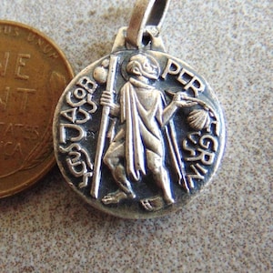 May include: A round silver-toned metal pendant featuring a relief of a figure holding a staff and a shell, with the text "S. JACOB PER GRIN" embossed around the edge, positioned next to a copper-colored one-cent coin for scale.