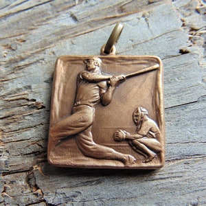 May include: Bronze-colored baseball pendant featuring a batter swinging at a pitch, with a catcher in the background. The square pendant has a loop at the top for a chain. The pendant is set against a weathered wood background.