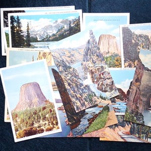 May include: A collection of vintage postcards featuring colorful landscape illustrations of American national parks, rock formations, canyons, and dams. The rectangular cards display various scenic views including Devils Tower, mountain ranges, and rivers.