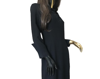 Könnte beinhalten: Schwarzes Kleid mit langen Ärmeln und knielangem Saum. Das Kleid hat eine Perlenverzierung am Ausschnitt und eine klassische, taillierte Silhouette. Das Kleid wird auf einer Schaufensterpuppe mit schwarzem und goldenem Gesicht präsentiert.