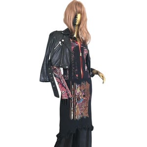 May include: A black leather jacket with silver zippers and a belt detail is worn over a black dress with a colorful graphic print. The dress has a ruffled hem. The mannequin has a gold arm and a painted face.