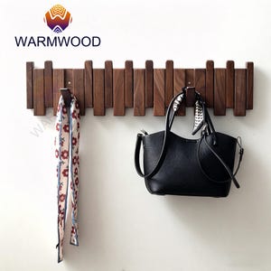May include: A wooden wall-mounted coat rack with a series of vertical wooden slats. A black handbag and a floral scarf hang from the hooks. The brand name "WARMWOOD" is displayed at the top left.