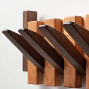 Peut inclure: Un porte-manteau mural composé d'une série de lattes en bois verticales aux tons brun clair et brun foncé alternés. Plusieurs patères en bois brun foncé s'étendent vers l'extérieur selon un angle ascendant, offrant des crochets de suspension.