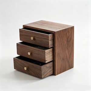 May include: A small wooden storage chest crafted from dark brown walnut wood, featuring three stacked drawers. Each drawer is equipped with a single, small, circular brass knob centred on the front panel. The wood grain is visible throughout.