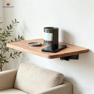 Op de afbeelding: Een rechthoekige, lichtbruine houten zwevende plank bevestigd aan de muur met zwarte metalen beugels. Op de plank staat een cilindrische metalen container met een wit label en een zwarte smartphone. De plank heeft een gladde afwerking.