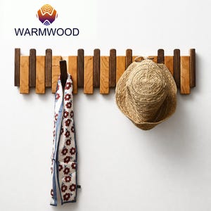 Könnte beinhalten: Eine Wandgarderobe aus Holz mit abwechselnd hellen und dunklen Holzlatten. Ein weißer Schal mit rotem Blumenmuster hängt an einem Haken, und ein beigefarbener, geflochtener Strohhut hängt an einem anderen. Das Logo WARMWOOD ist oben zu sehen.