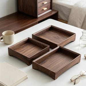 May include: Three rectangular wooden trays crafted from dark brown walnut wood with visible grain patterns. Each tray features raised edges and small feet at the corners, providing a stable base for organizing items on a flat surface.