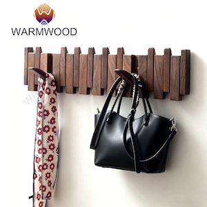 May include: A dark brown wooden coat rack with a distinctive design, mounted on a wall. The rack features multiple hooks, holding a black handbag and a floral scarf. The brand name "WARMWOOD" is displayed above the rack in the image.