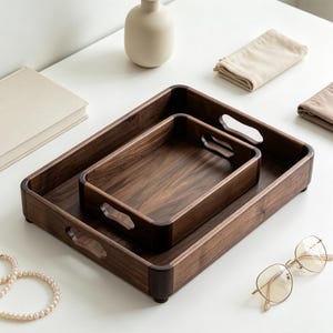 May include: Two rectangular dark brown wooden serving trays with rounded corners and integrated cutout handles. The trays are nested, featuring a visible wood grain pattern, smooth edges, and small feet on the base for stability on flat surfaces.