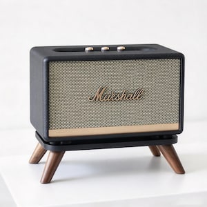 May include: A black Marshall speaker with a textured beige grille, gold-toned trim, and three top-mounted control knobs. The rectangular speaker is elevated on a black platform supported by four angled, dark brown wooden legs.