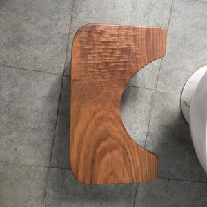 May include: A rectangular wooden bathroom stool with a curved cutout along one side, featuring a rich brown wood grain pattern with visible natural swirls and a smooth, polished finish, viewed from directly above in a top-down perspective.