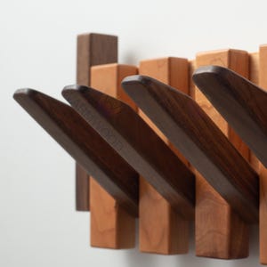 May include: Wooden wall-mounted coat rack featuring a series of angled hooks. The hooks are a dark brown, contrasting with the lighter wood of the base. The design is modern and minimalist, suitable for entryways or bedrooms.