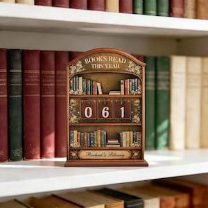 May include: A wooden bookshelf-shaped counter with the text "BOOKS READ THIS YEAR" and the numbers "061" displayed. The counter is decorated with floral accents and the words "Rachael's Library". The background features a shelf of books.